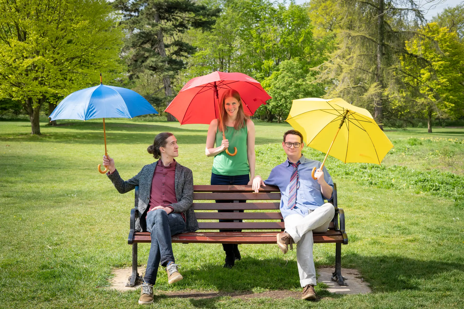 Two men and a woman on a park bench, holding red, blue and yellow umbrellas